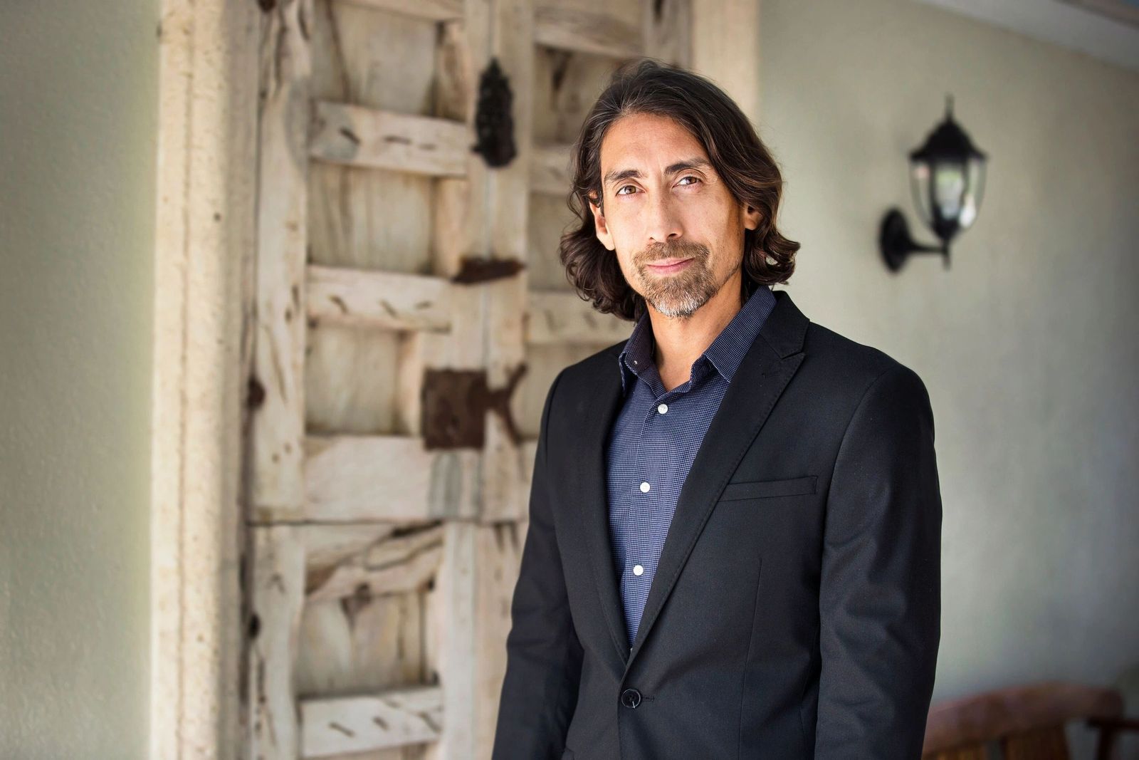 Sayer Ji in a dark blazer and blue shirt, standing beside a weathered wooden door
