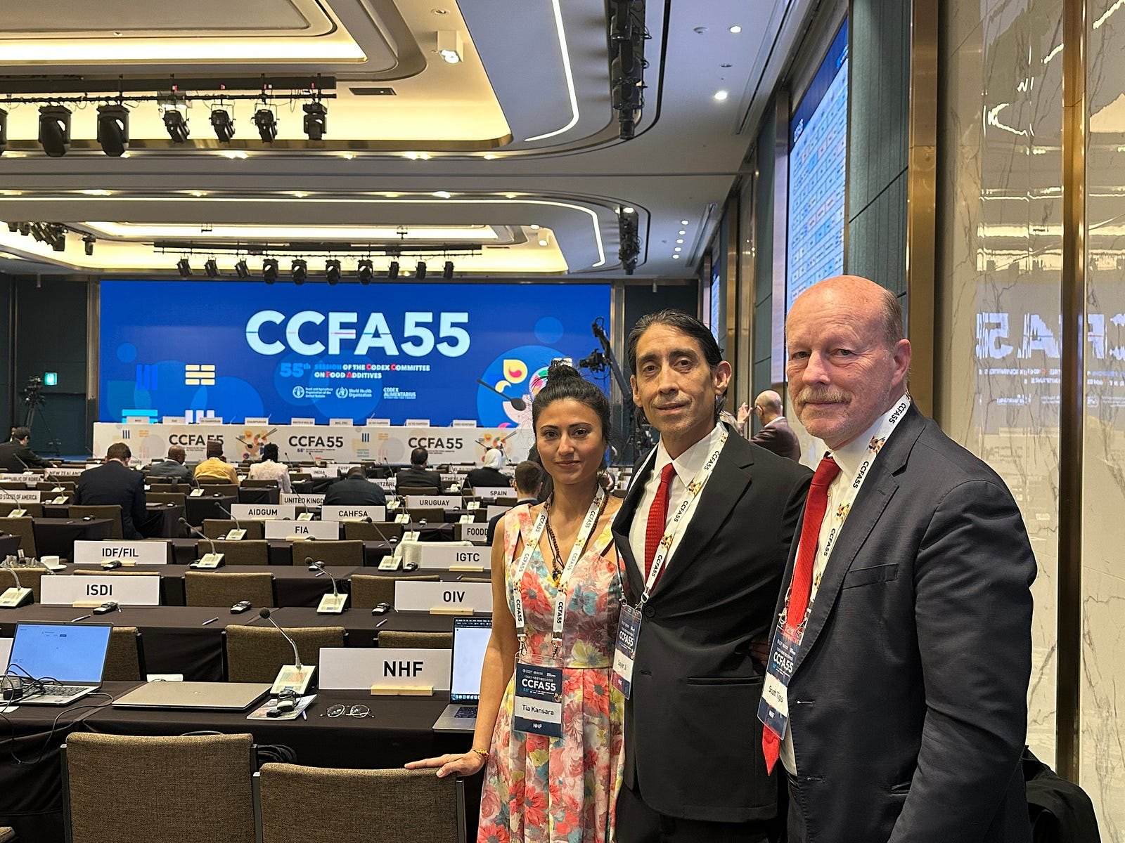 Dr. Tia Kansara, Sayer Ji and Scott Tips wearing CCFA55 delegate badges in the Codex meeting hall in Seoul, an NHF placard on the desk beside them and the CCFA55 screen behind.