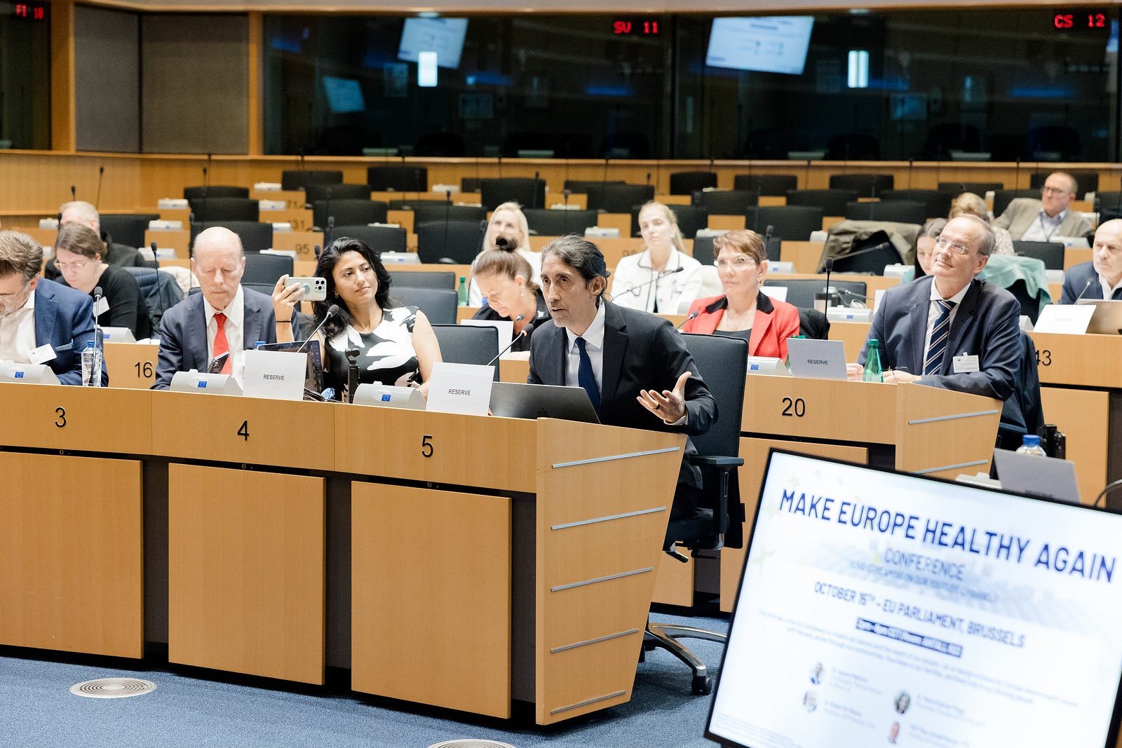Sayer Ji speaking from a desk in a European Parliament committee room, one hand raised, with a screen in the foreground reading Make Europe Healthy Again, October 15th, EU Parliament, Brussels.