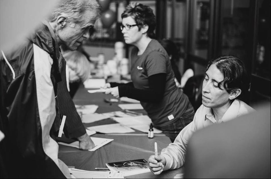Sayer Ji seated at a signing table, pen in hand, talking with attendees after a speaking event